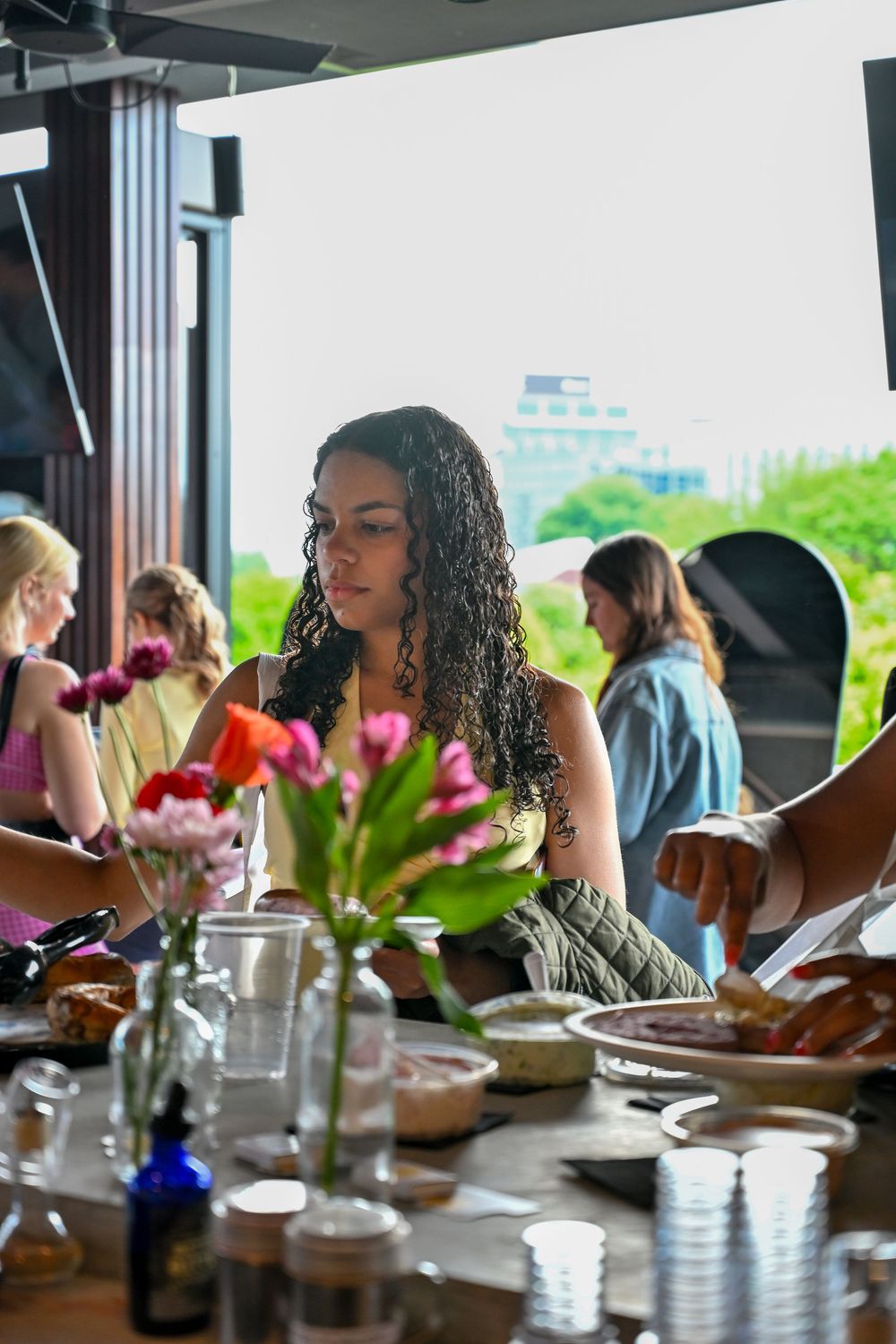 Young woman with curly hair at a buffet table with flowers, surrounded by guests at an outdoor event.