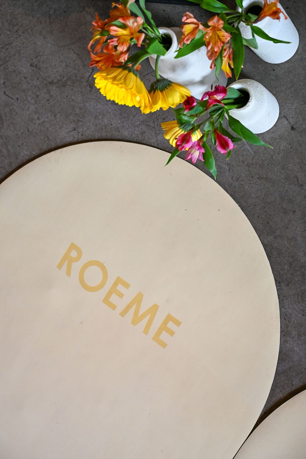 Round beige ROEME branded menu beside colorful floral arrangement in white vases on gray surface.