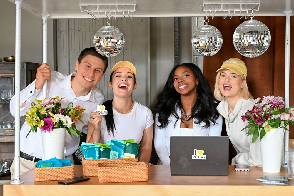 Four smiling friends pose behind a bar counter with flowers, disco balls, and a laptop displaying a logo.