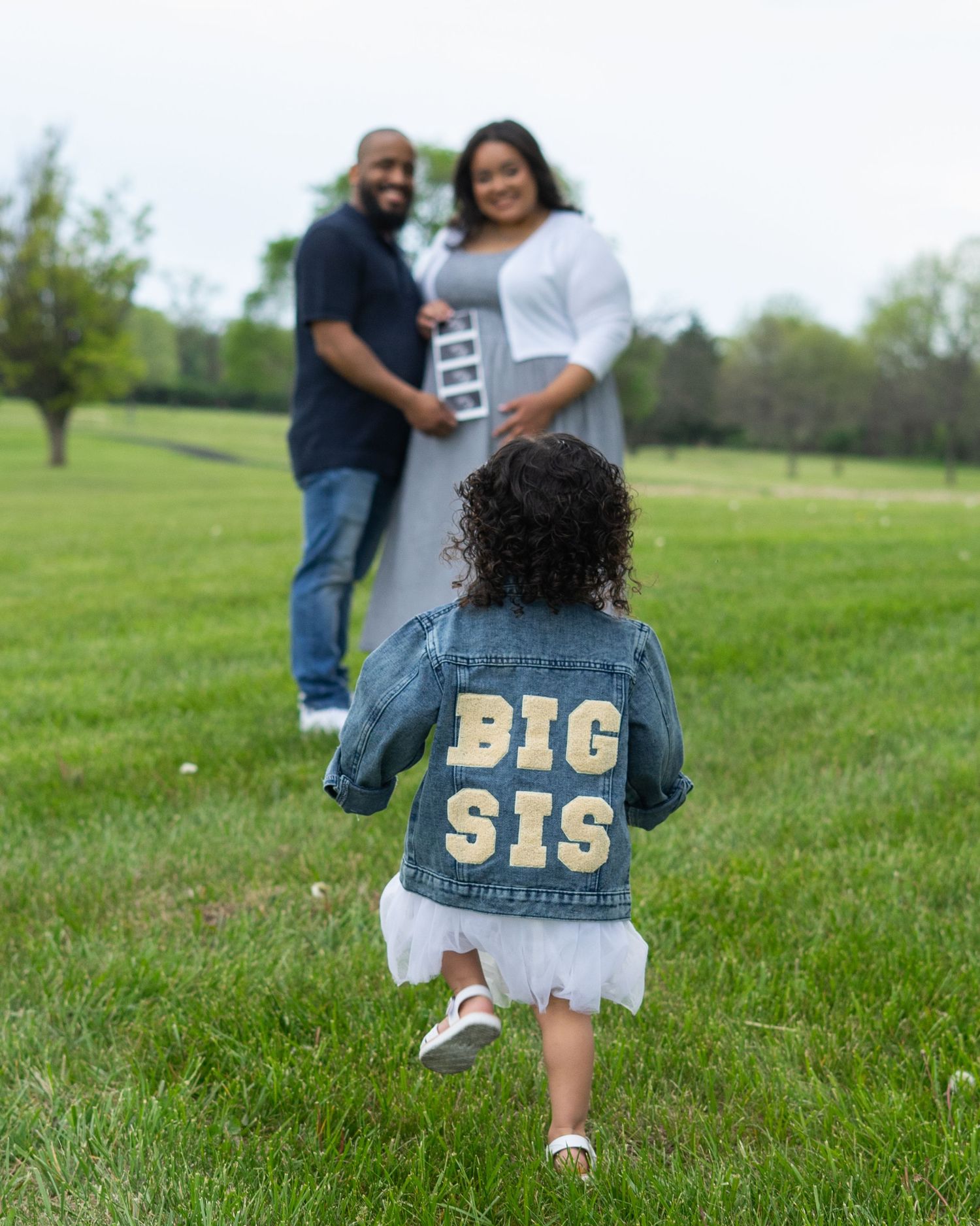 A toddler in a denim Big Sis jacket runs toward her expecting parents on a green lawn.