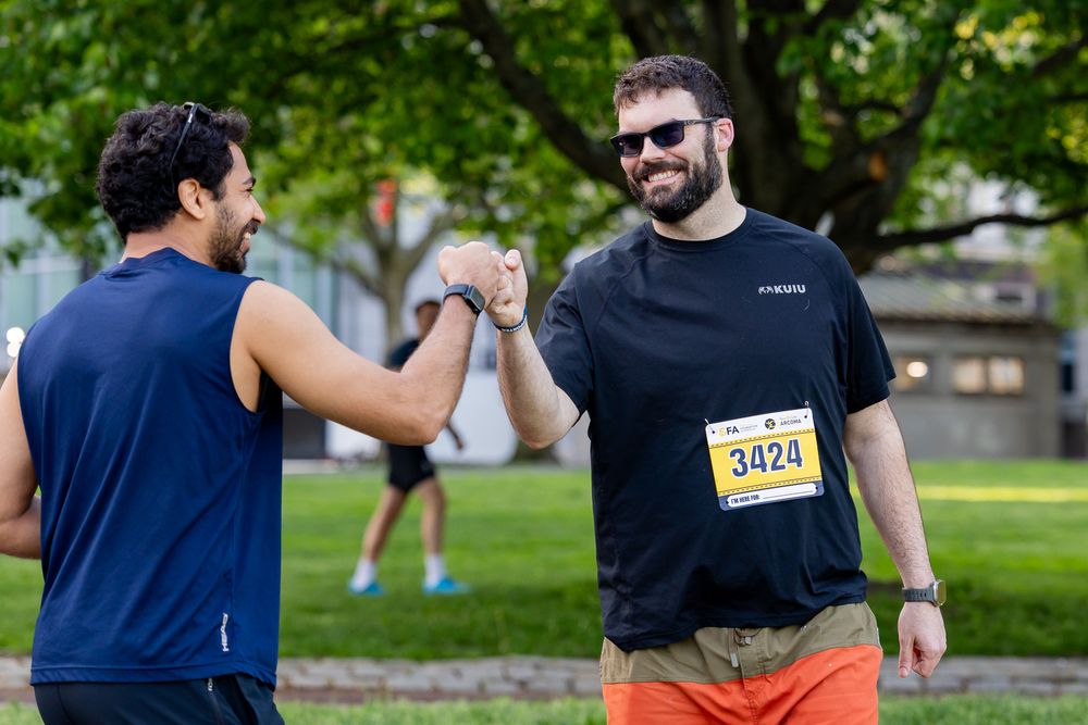 Two men fist bumping at an outdoor race event, one wearing a race bib number 3424, smiling in a park.