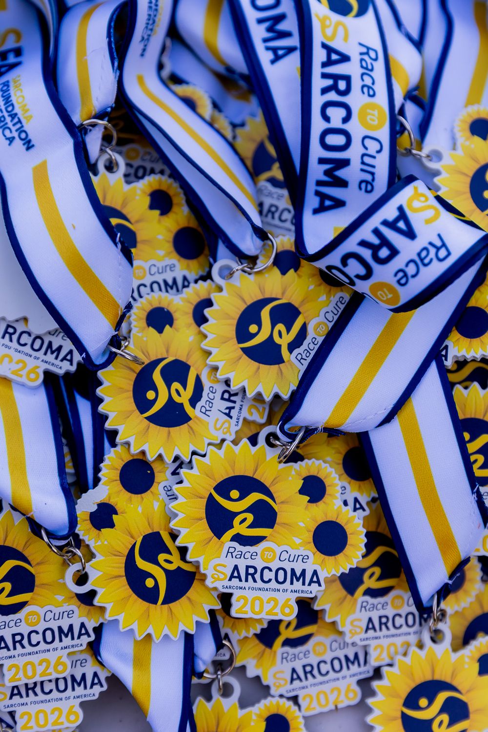 Pile of Race to Cure Sarcoma 2026 medals with blue and white ribbons and yellow sunflower designs.