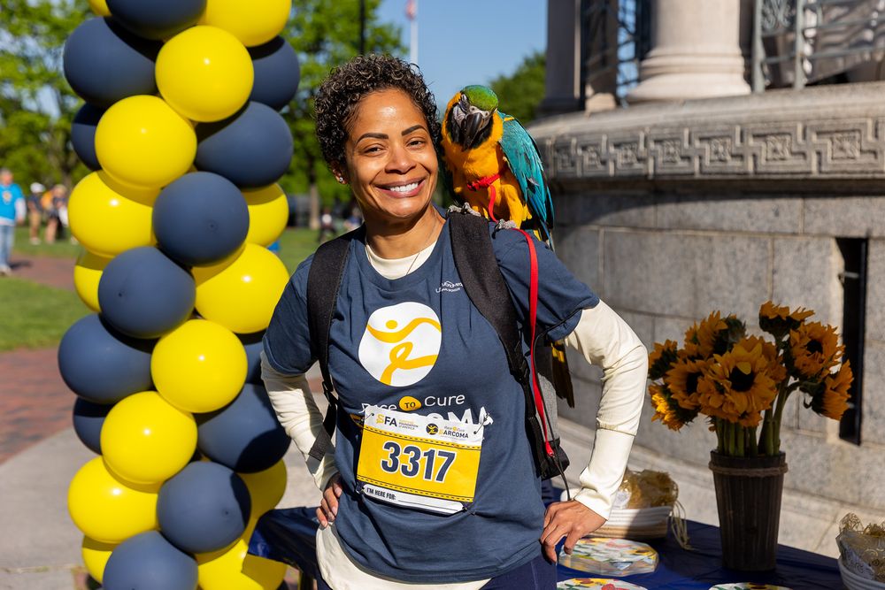 Woman with parrot on shoulder wearing race bib 3317 smiling at charity run event with balloon column.