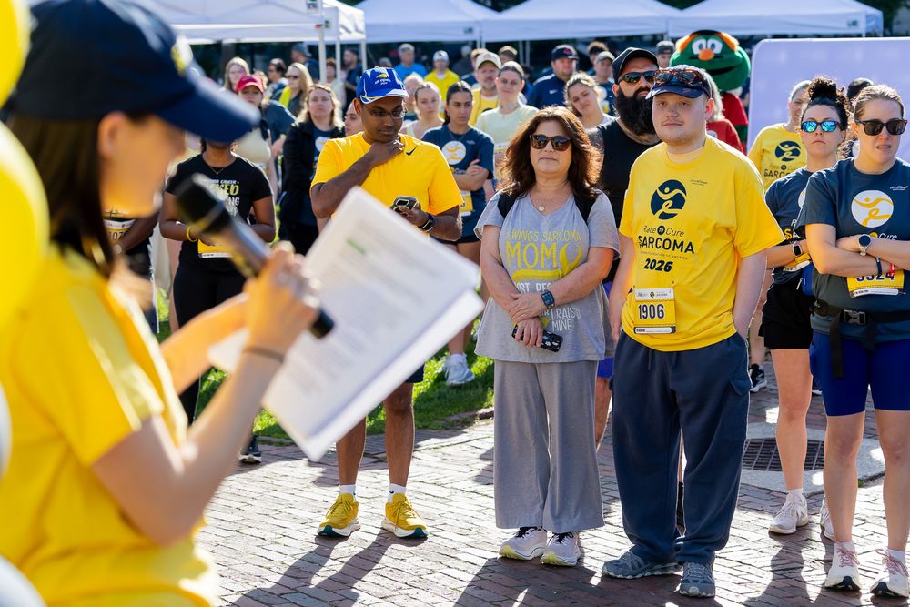 Crowd of participants in blue and yellow attire listen to a speaker at an outdoor charity walk event.