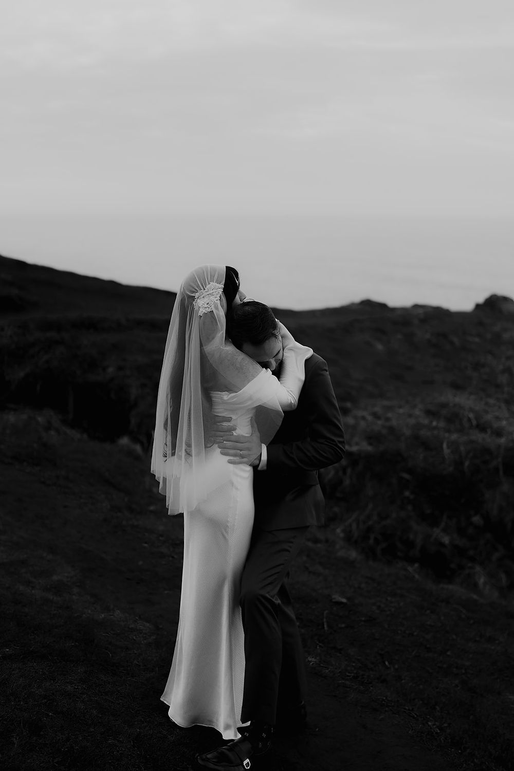 Black and white photo of a bride and groom embracing on a rocky hillside, her veil flowing as they share a romantic moment.