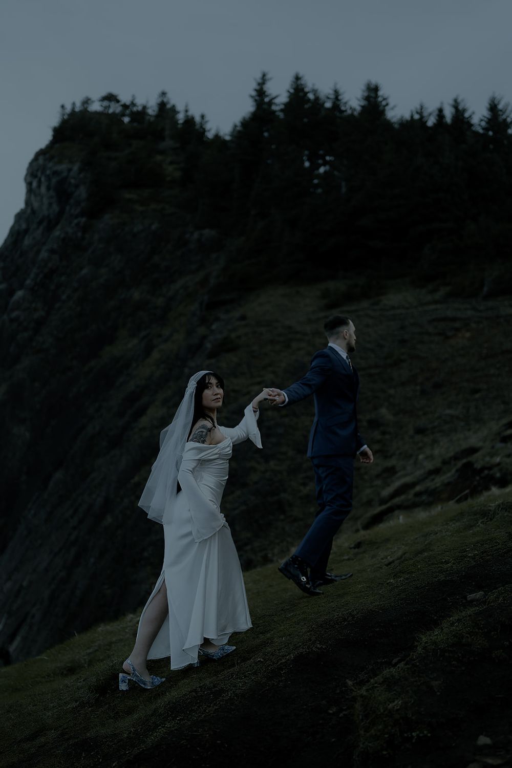 Bride and groom dancing on a misty mountain slope at dusk, wearing wedding attire with evergreen trees in background.