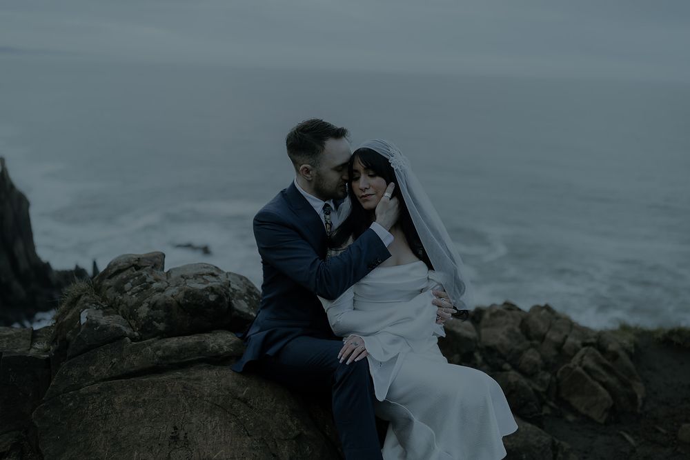 Couple in wedding attire embracing on rocky coastal cliffs with moody ocean waves in the background.