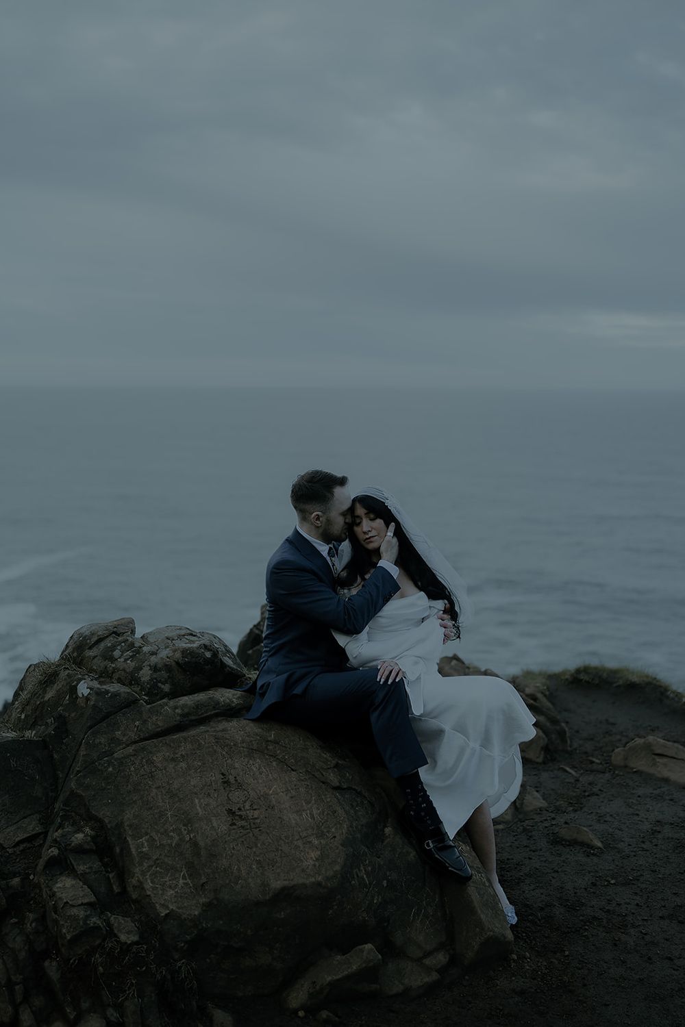 Couple in wedding attire sitting on coastal rocks, sharing a tender moment with moody ocean backdrop.