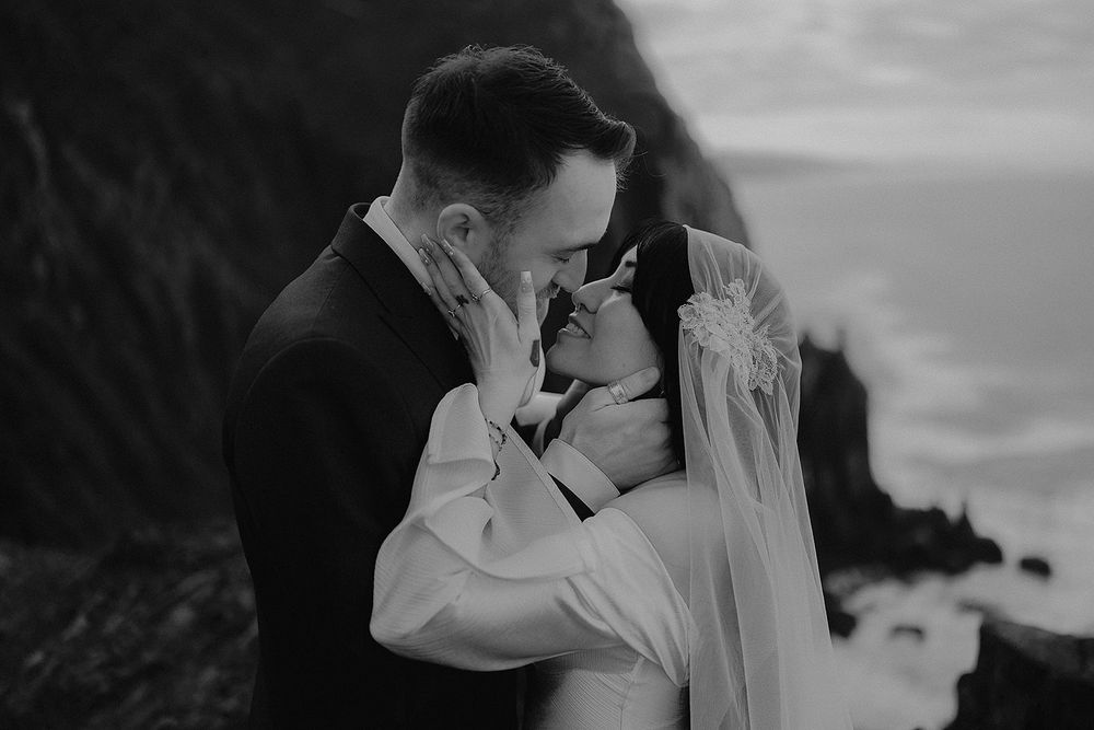 Black and white photo of bride and groom embracing near dramatic coastal cliffs on wedding day.