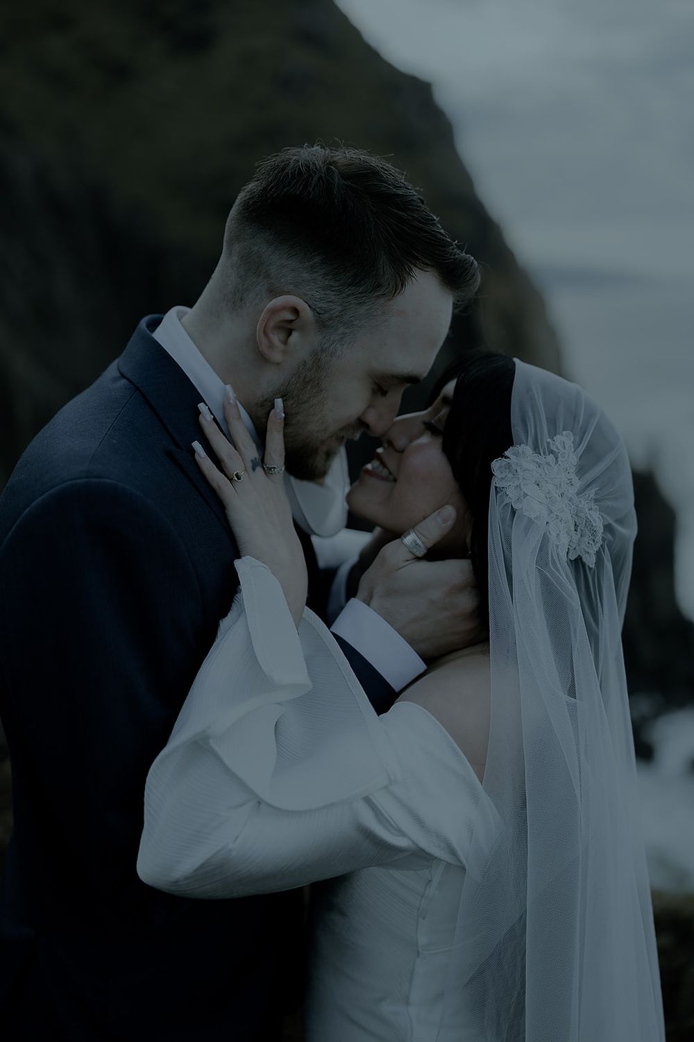 Bride and groom share an intimate moment outdoors, with rocky cliffs and ocean in the background.