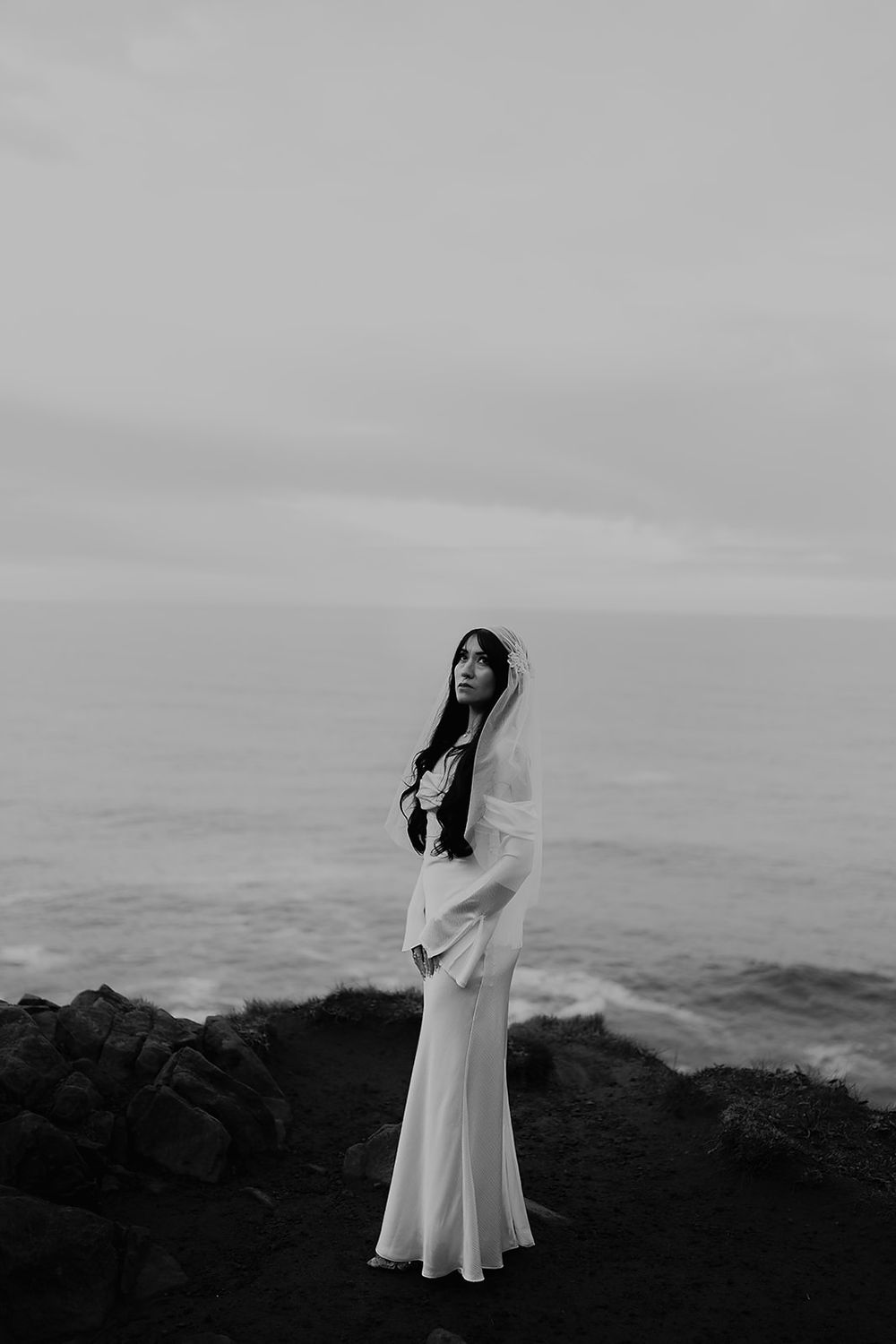 Black and white photo of a bride in a white gown and veil standing on rocky coastal cliffs with ocean waves behind her.