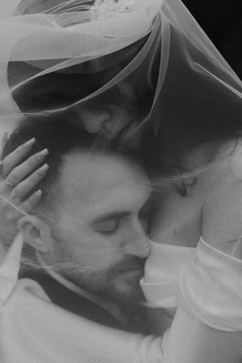 Black and white wedding photo of bride and groom sharing intimate moment under flowing veil.