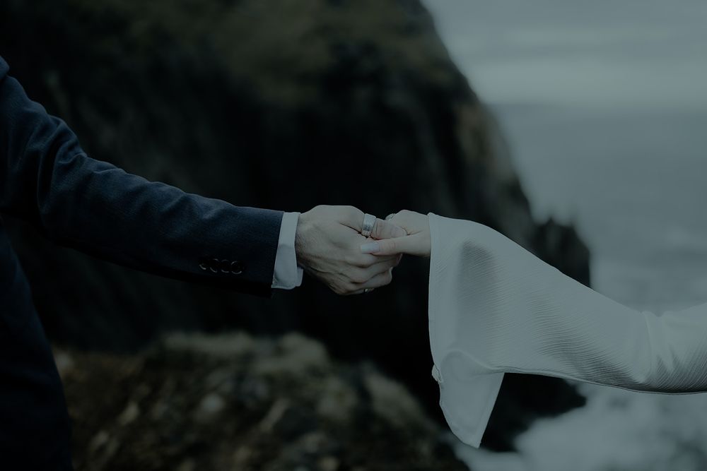 A groom in a navy suit holds the hand of a bride in a white dress on rocky coastal cliffs.