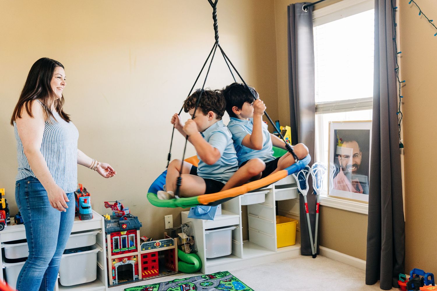 Two boys swing on an indoor hanging swing in a colorful playroom while their mom watches and smiles nearby.