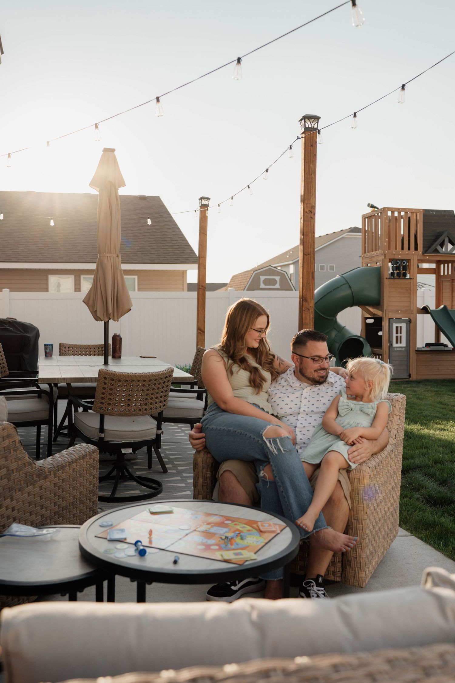 Family of three relaxing on patio furniture in backyard at sunset with string lights and playground.
