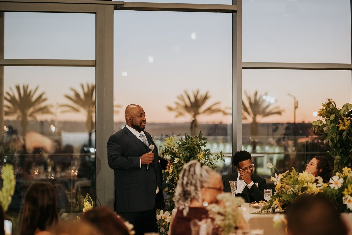 Man giving a wedding toast at an elegant reception venue with palm trees and sunset views through large windows at The Lane.