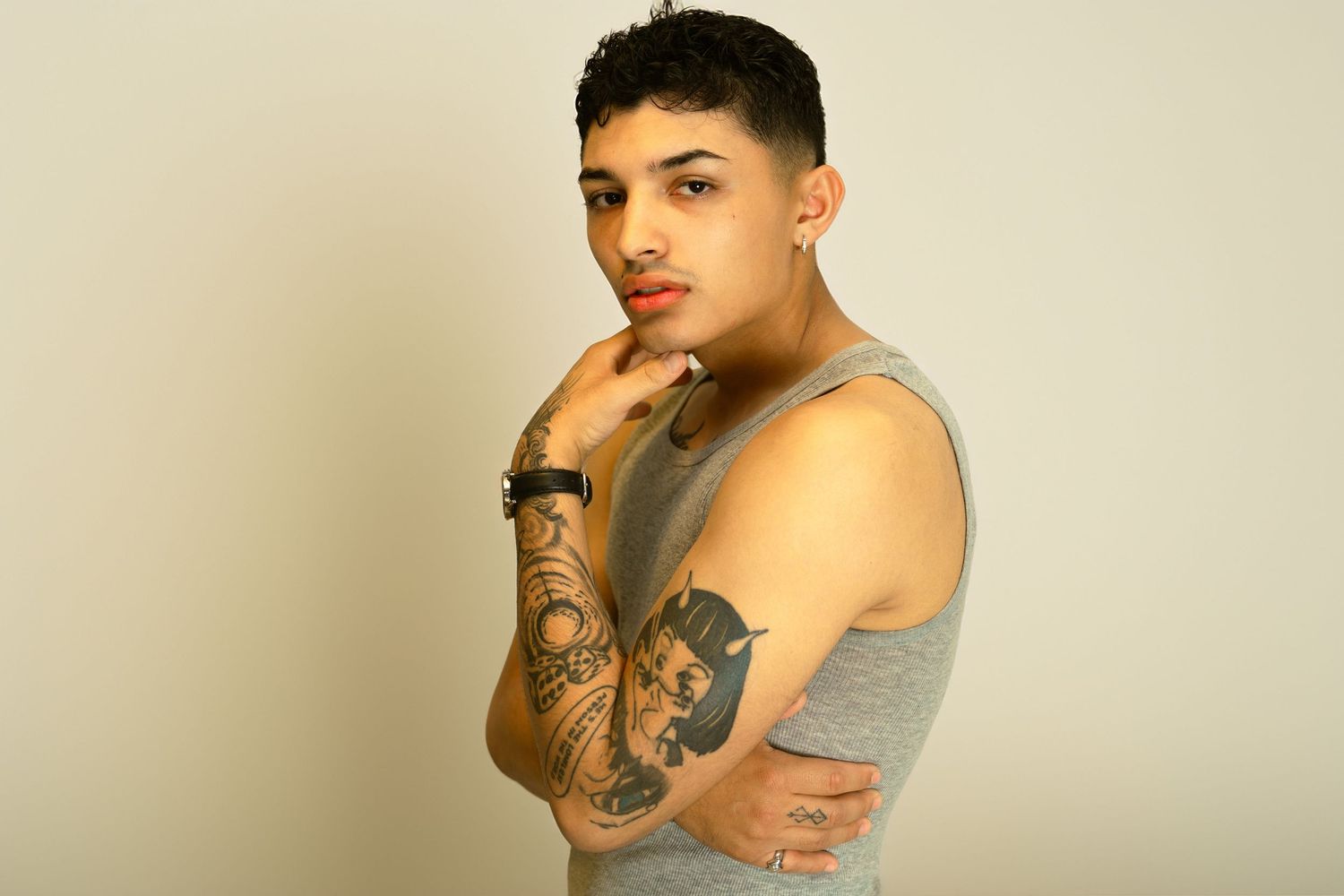 Young man with tattoos on arms wearing gray tank top, posing with hand on chin against light background.