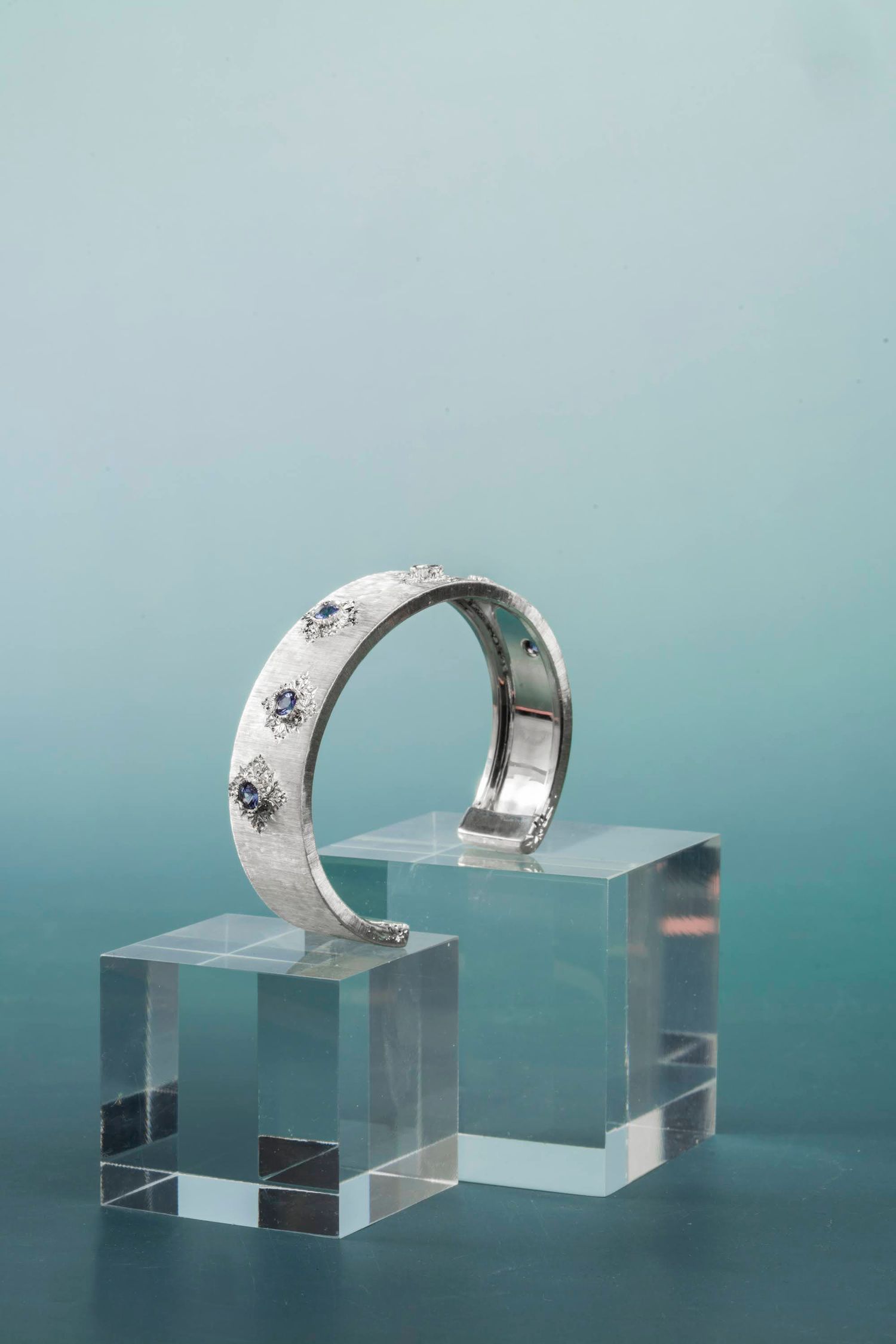 Silver cuff bracelet with blue gemstones displayed on clear acrylic blocks against a teal background.