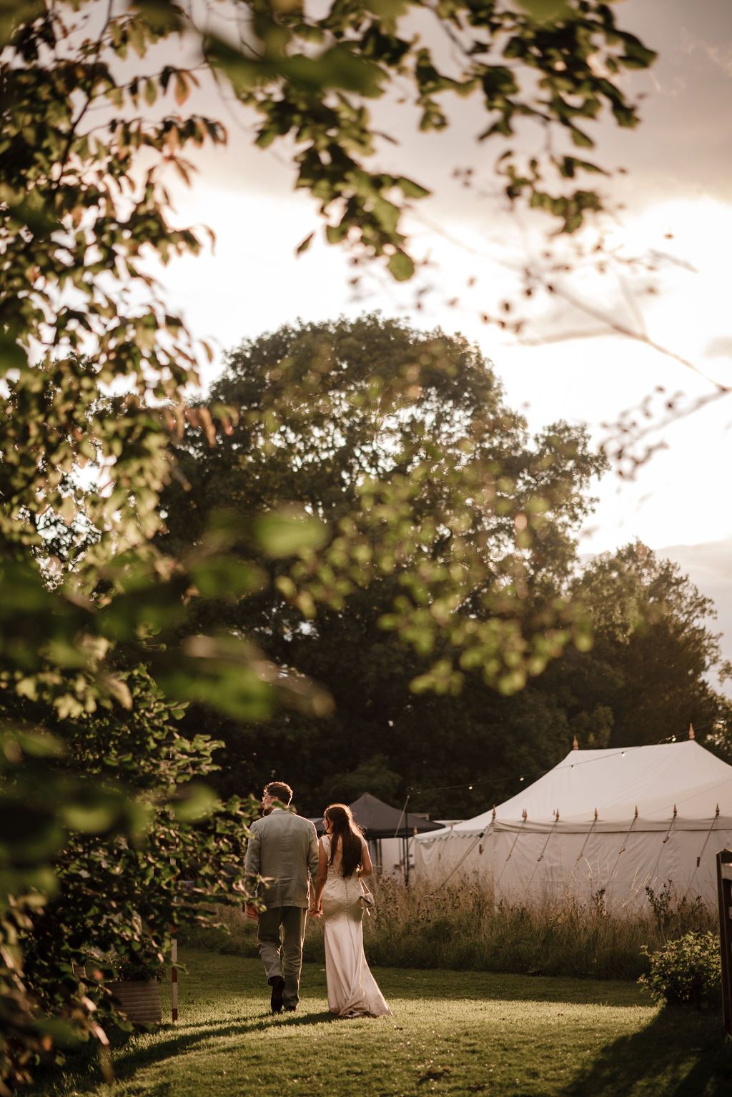 Couple walking hand in hand through lush garden toward elegant white marquee tent at golden hour wedding reception.