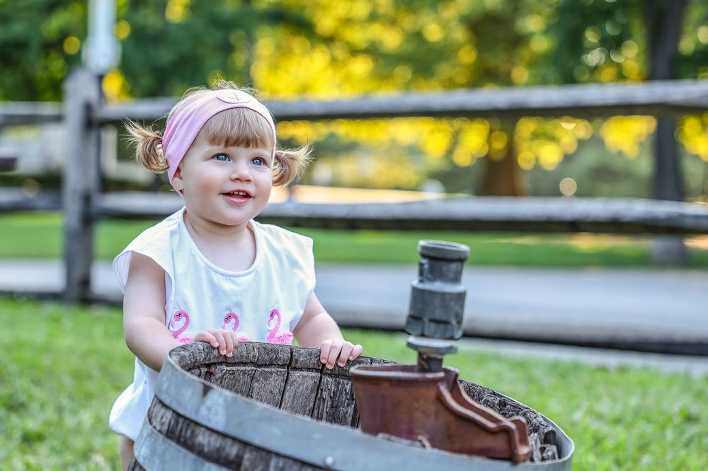Happy toddler girl in white shirt playing in wooden barrel outdoors.