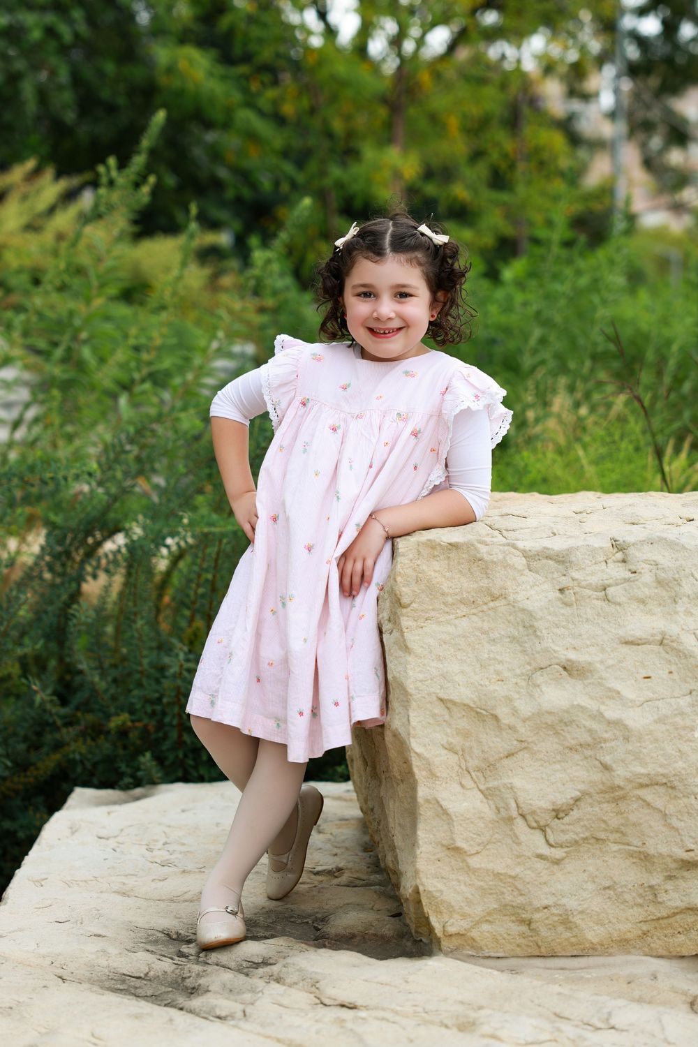 A smiling young girl in a pink dress and white top leans against a large rock outdoors, surrounded by green trees.
