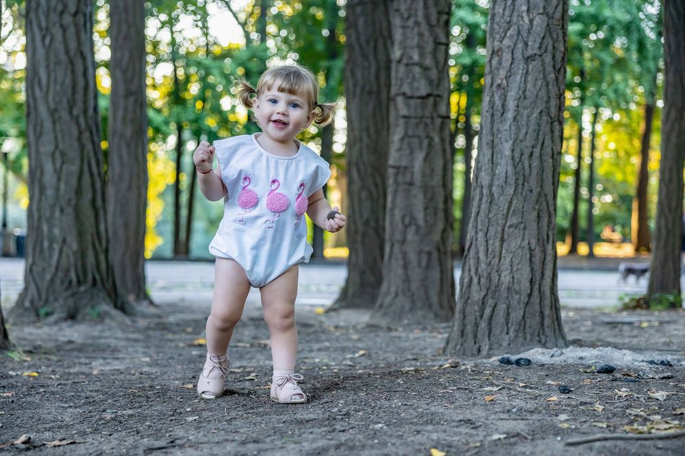 Toddler girl in flamingo romper walking among trees in sunny park.