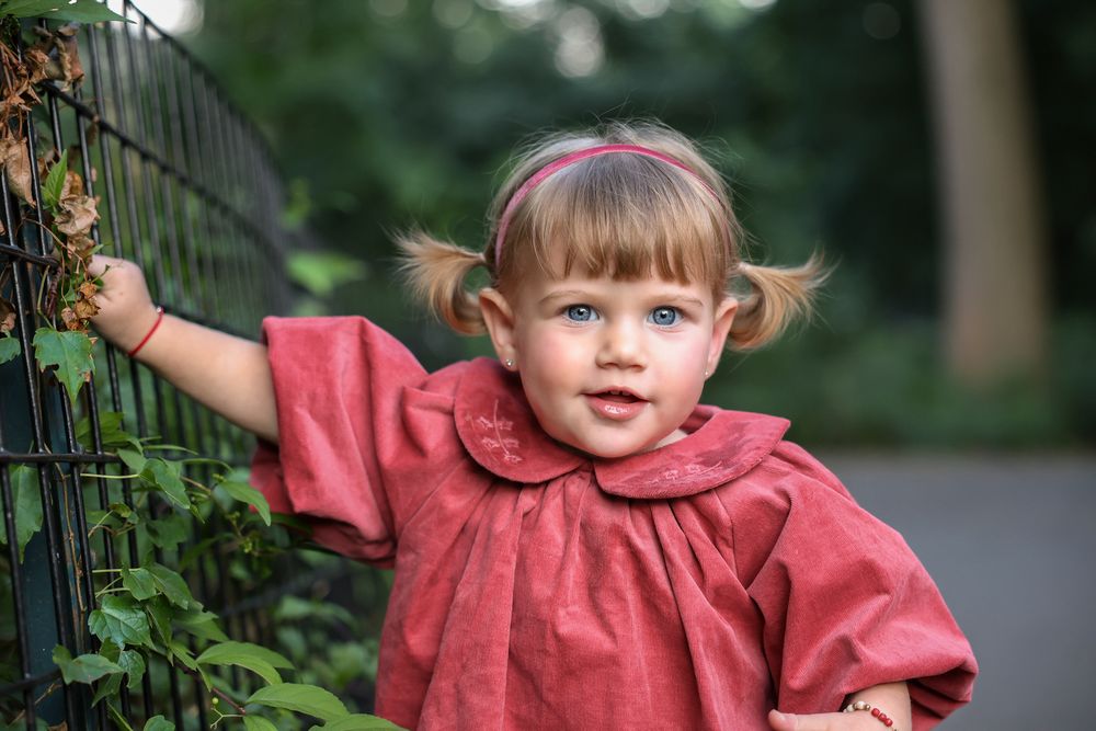 Blue-eyed toddler girl in red dress leaning against a metal fence surrounded by greenery.
