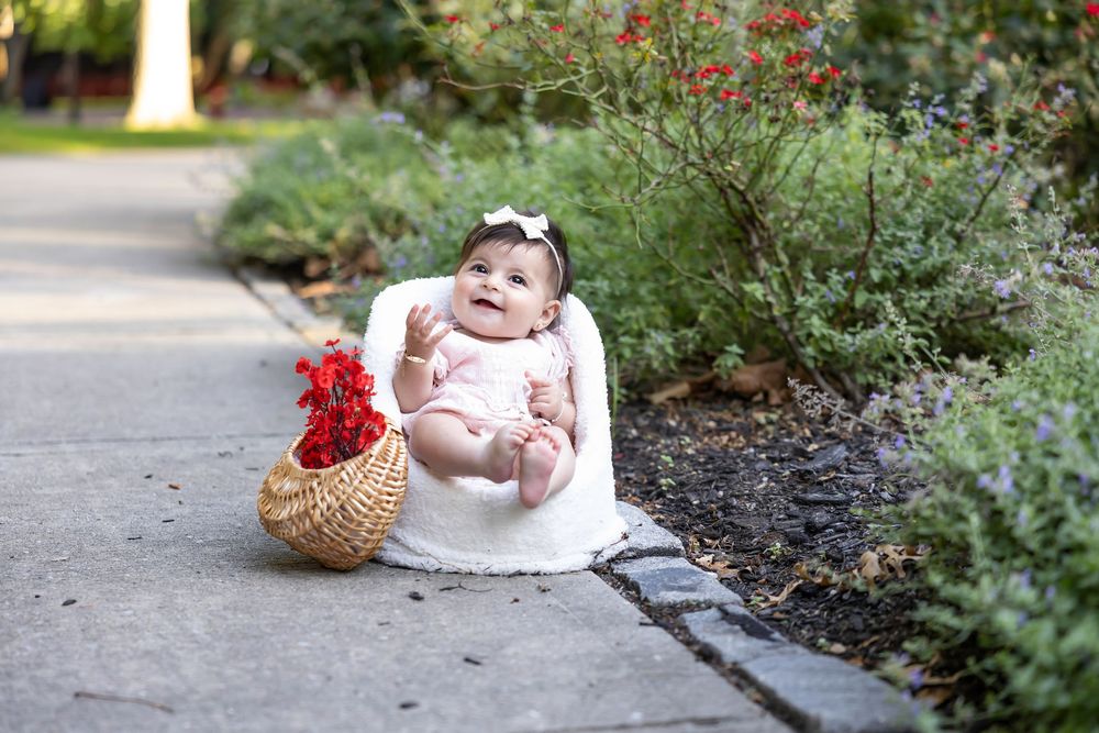 Sweet baby girl in white dress sitting in a chair on a garden path with a flower basket.