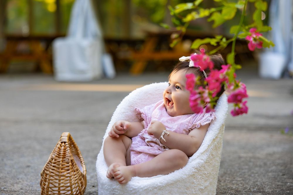 Laughing baby in pink outfit sitting in white chair near pink flowers.
