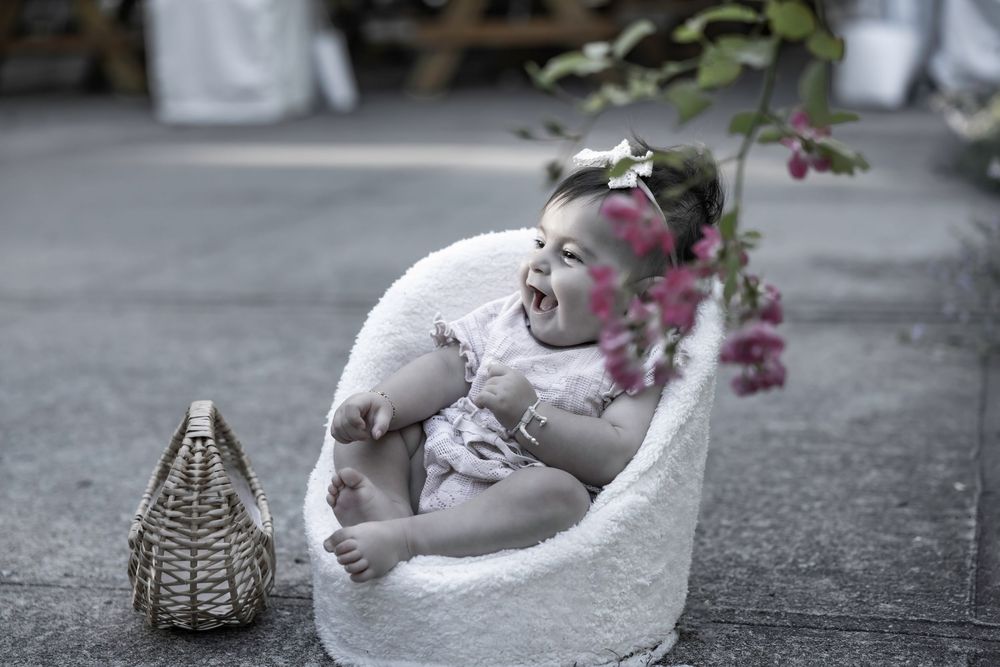 Laughing baby resting in a white fluffy chair surrounded by pink flowers and a wicker basket.