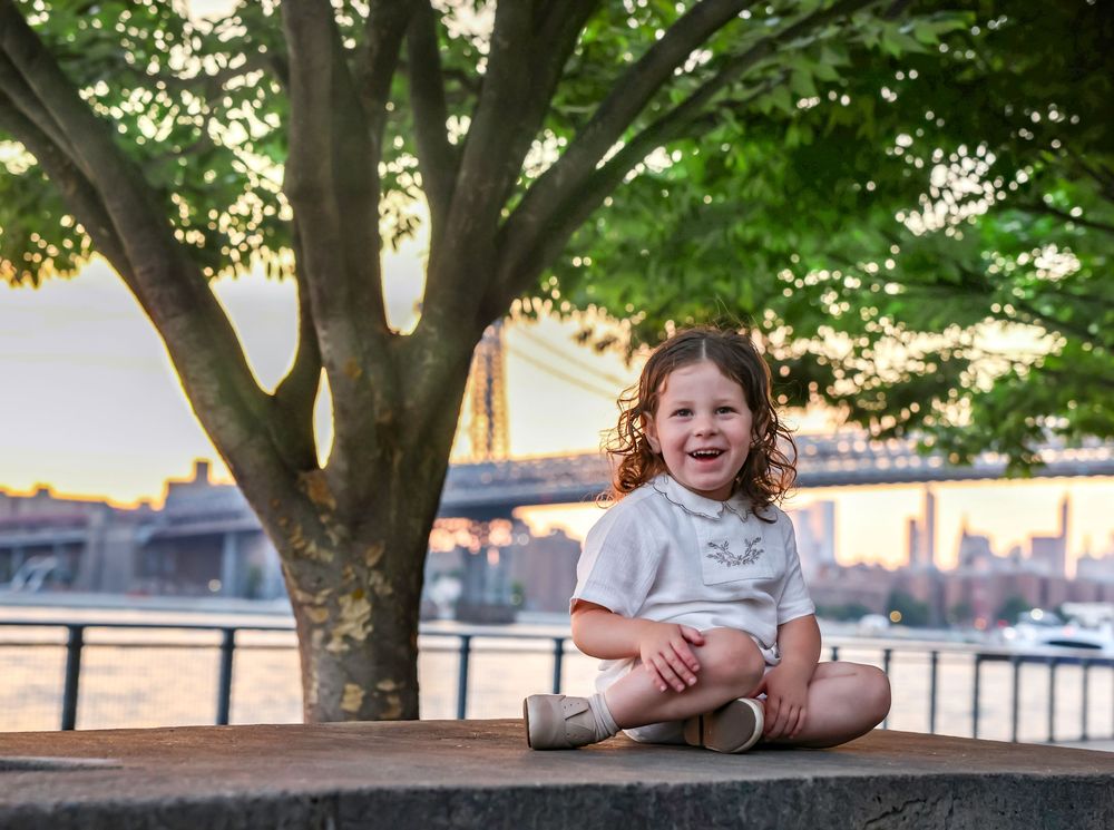 Happy toddler girl with curly hair sitting on stone wall laughing with Brooklyn Bridge in background.