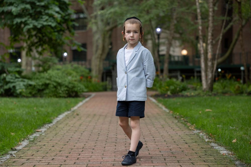Young boy in light blazer and shorts walking confidently on brick path in urban park setting.