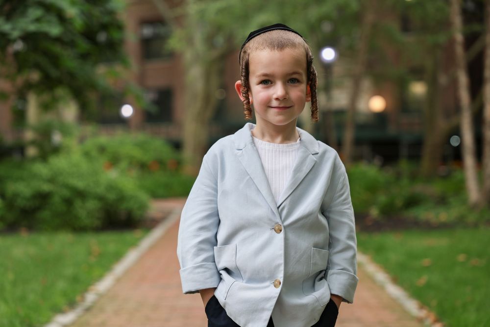 Smiling young boy in light gray blazer standing on brick pathway in a lush green park.