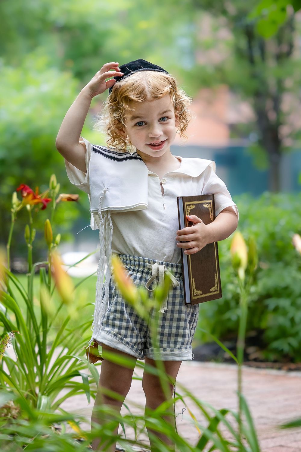 Curly-haired toddler boy in sailor outfit holding a book and wearing a kippah in a garden.