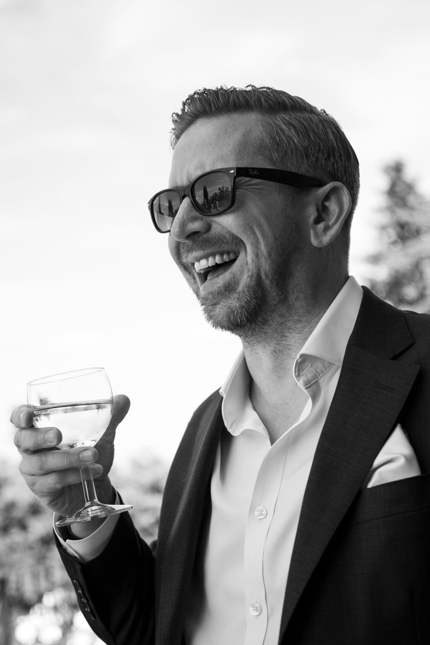 Smiling man in suit and sunglasses holding wine glass outdoors in black and white photo.