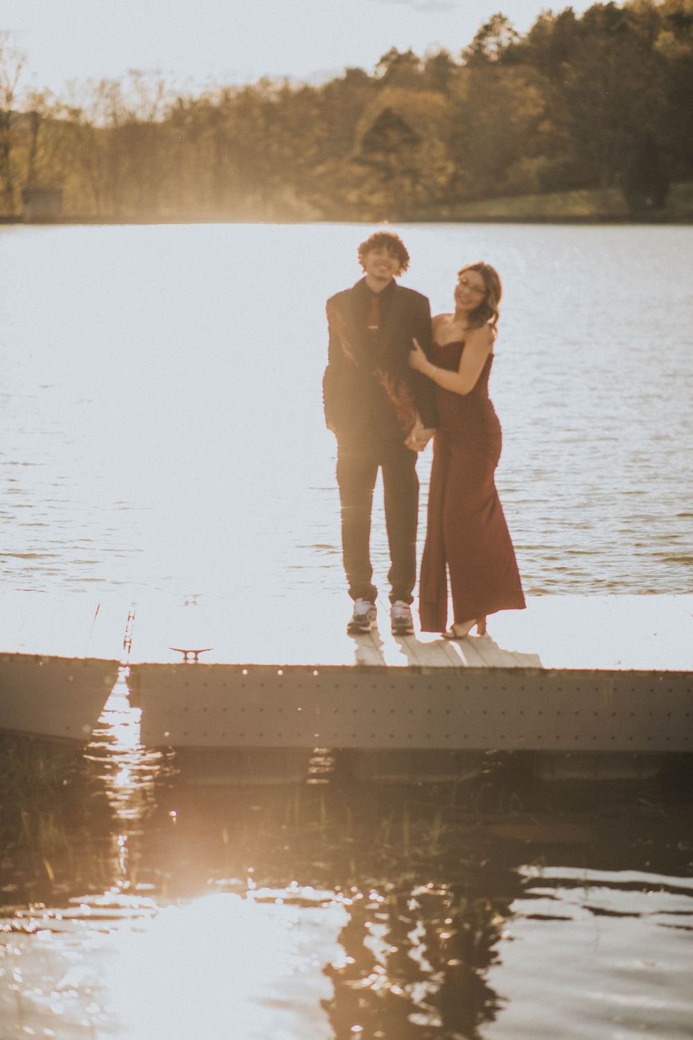 A couple in formal attire stands on a wooden dock by a lake, the woman in a red gown and man in black suit. indiana pa photographer prom