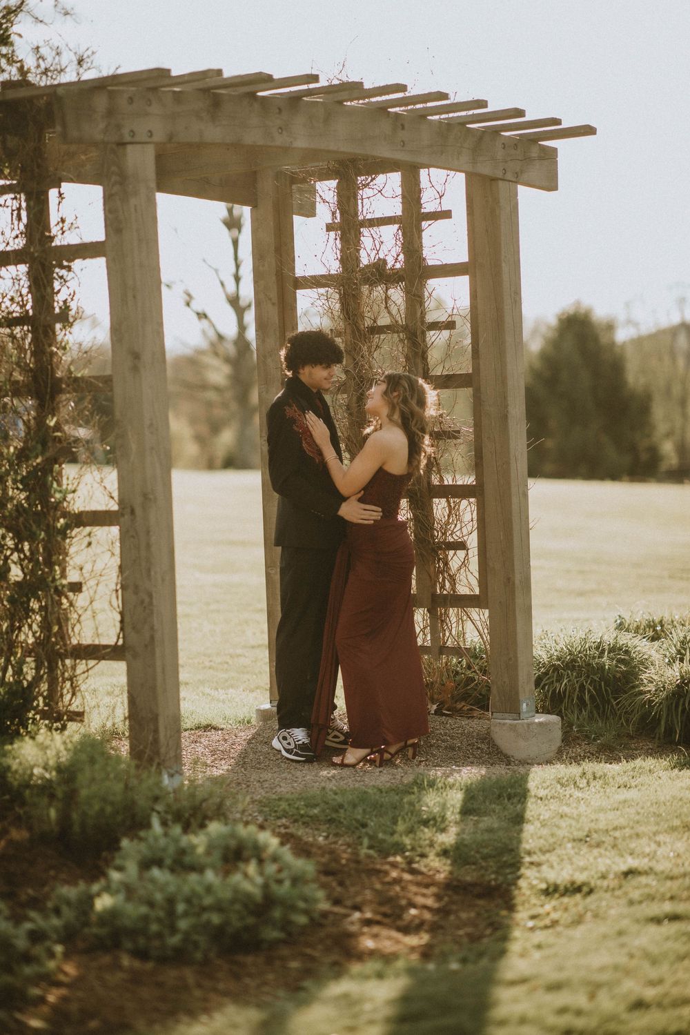 Couple embracing under a wooden pergola in a lush garden, wearing formal attire for a romantic outdoor photo. indiana pa photographer senior