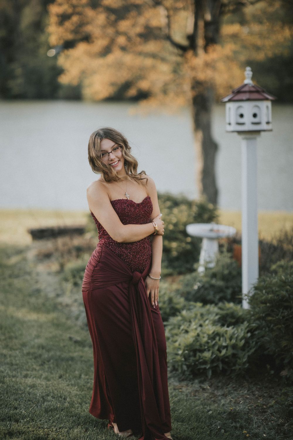Young woman in elegant strapless burgundy gown posing outdoors near a birdhouse indiana pa photographer, senior in prom dress