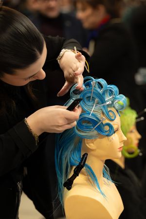 Une apprentie utilise des pinces pour peaufiner une perruque bleue aux boucles élaborées sur une tête de mannequin. MAF 2026