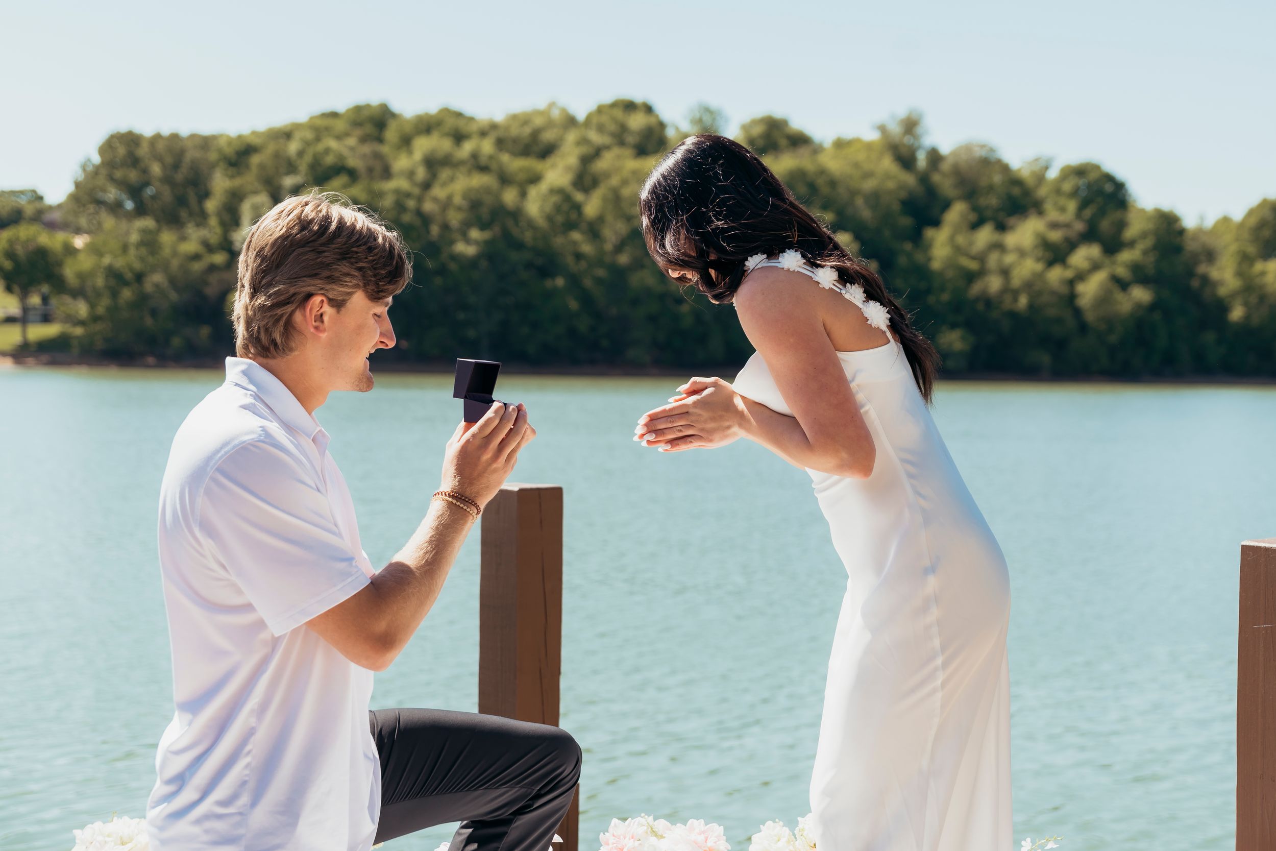 Man kneeling on a dock proposing to a woman in a white dress by a serene blue lake.
