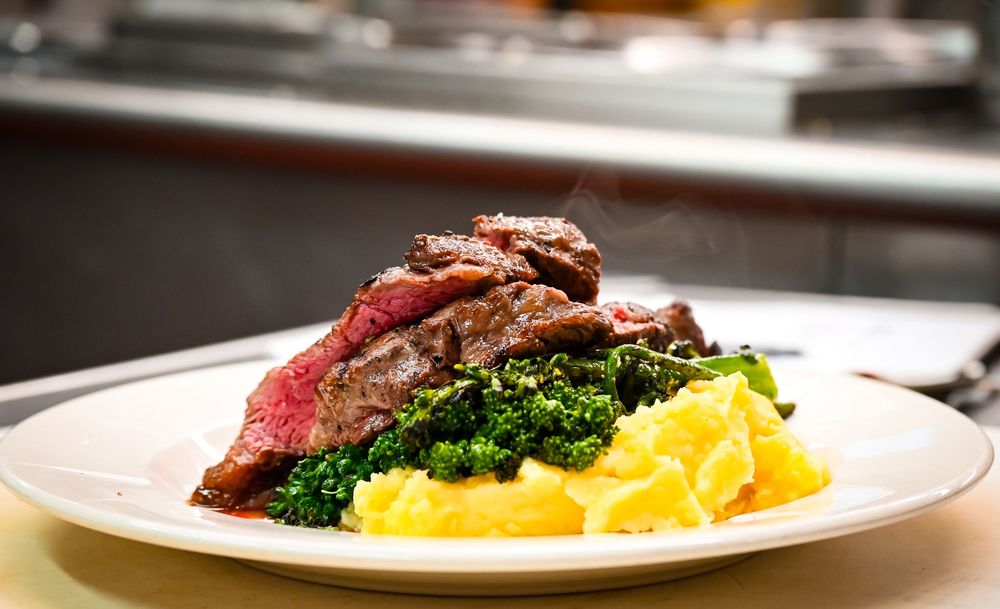 Juicy sliced steak served over creamy mashed potatoes with sautéed kale on a white plate in a restaurant.