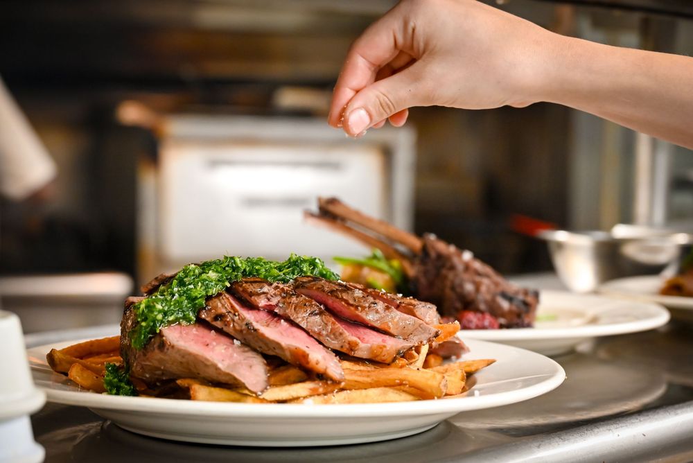 Chef seasoning perfectly sliced medium-rare steak with herb topping served on a plate in a professional kitchen.