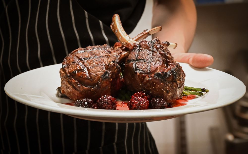 Grilled lamb chops served on a white plate with fresh blackberries and asparagus, held by a chef in striped apron.