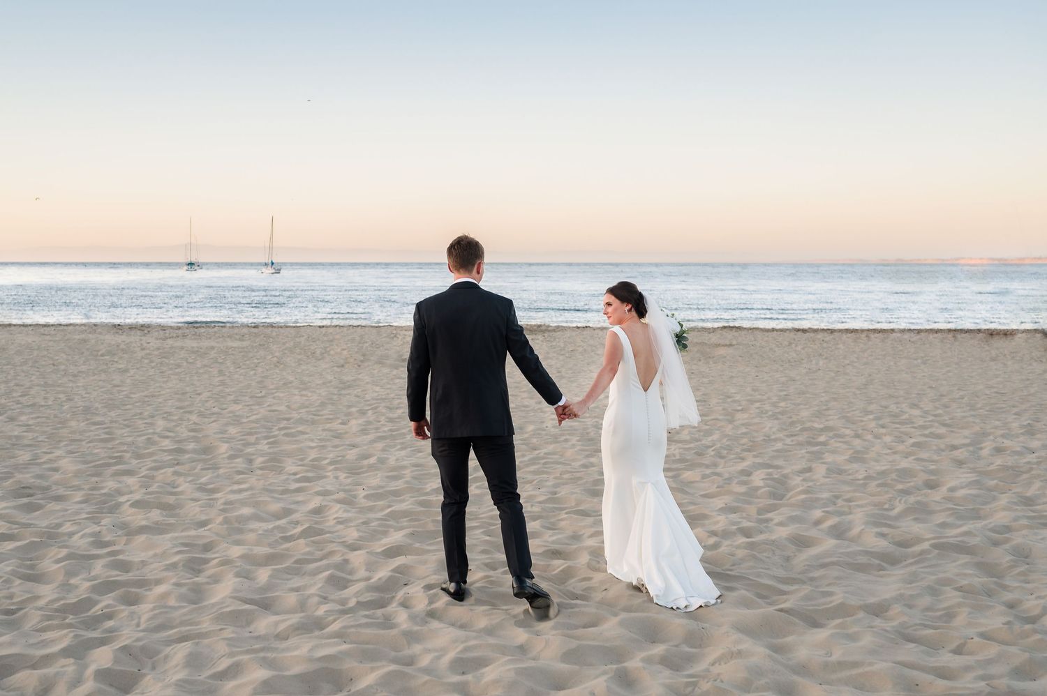 Wedding party celebrates with champagne by the ocean, bride in white gown, groom in tan suit, bridesmaid in sage green dress.