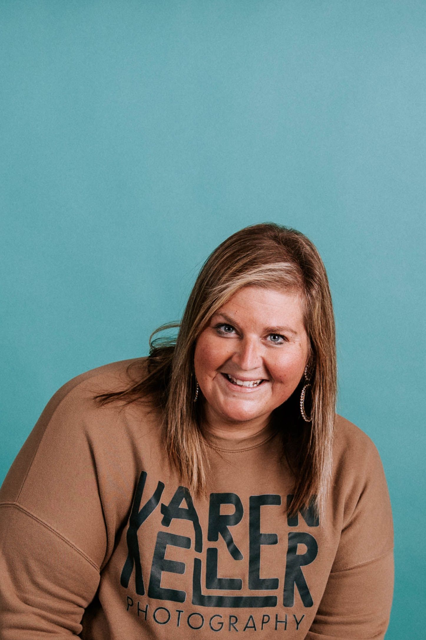Woman smiling in a tan Karen Keller Photography sweatshirt against a teal background.
