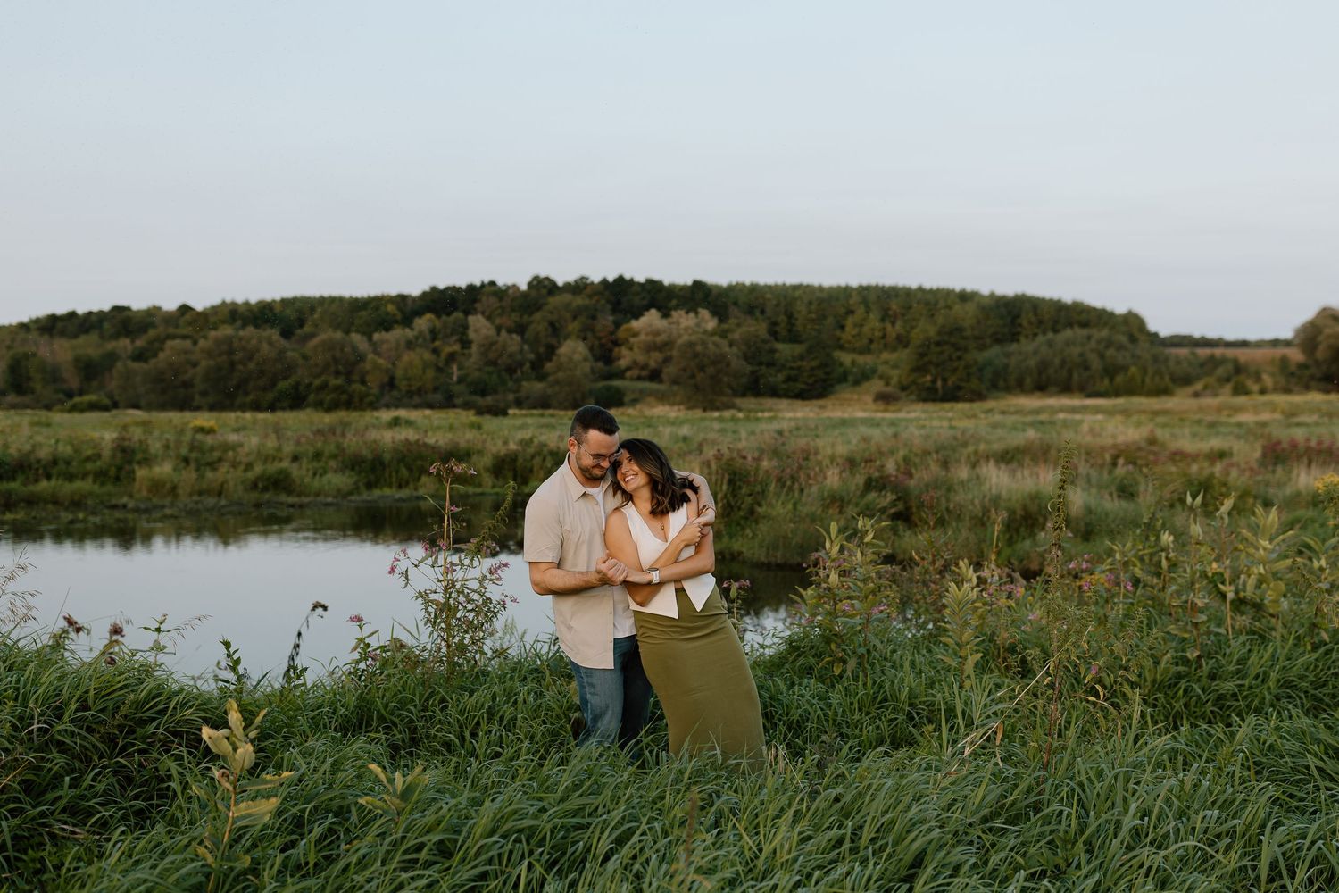 Couple embracing near a serene pond surrounded by lush greenery and trees at dusk in a romantic outdoor setting.