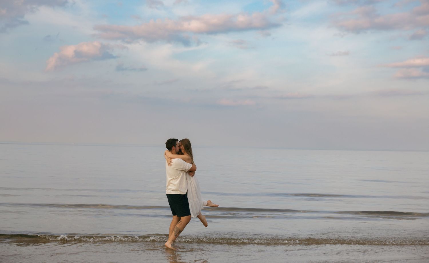 Couple embracing and kissing on a serene beach at dusk with soft pastel clouds over calm ocean waters.