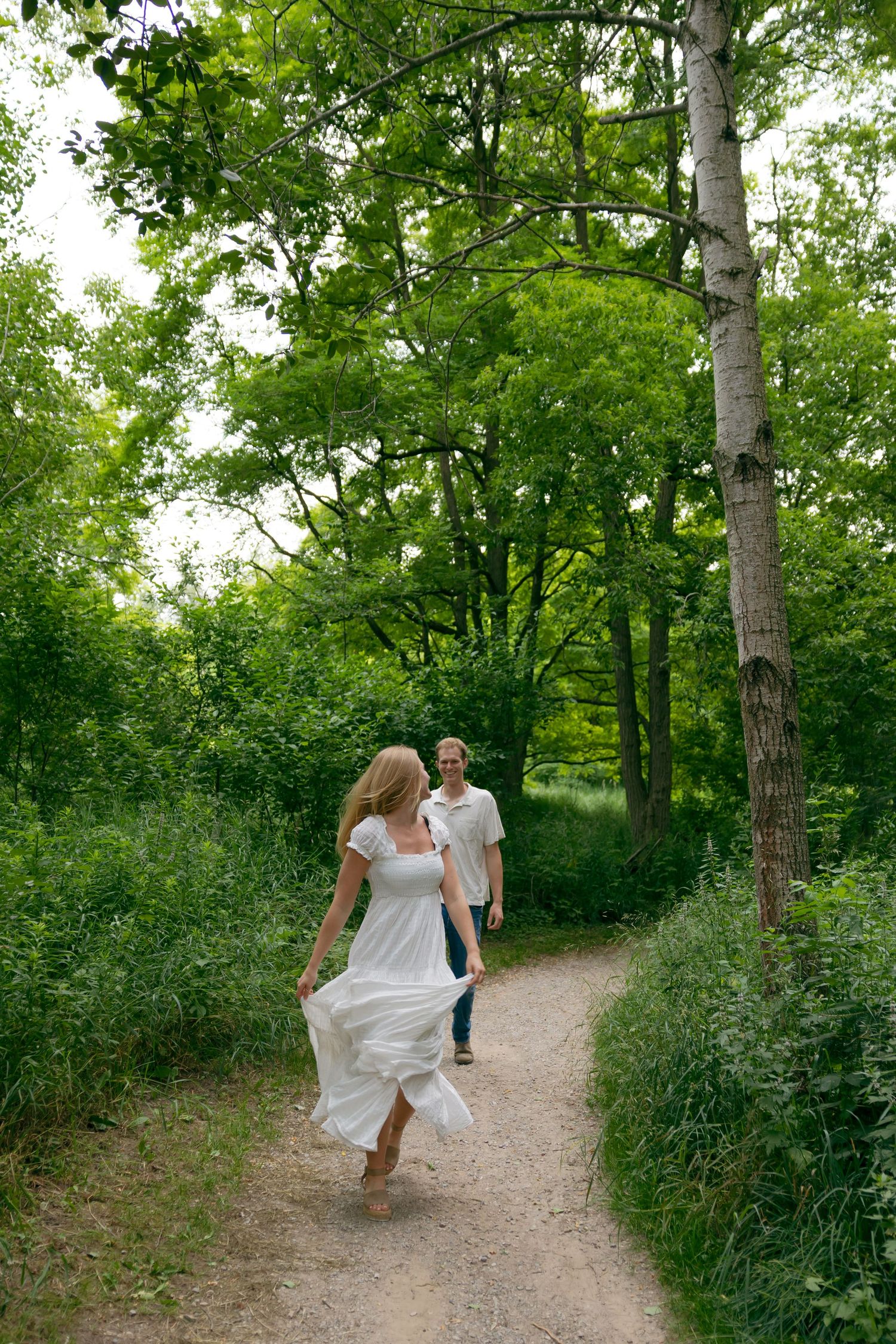 A woman in a white dress runs toward a smiling man on a lush green forest path.