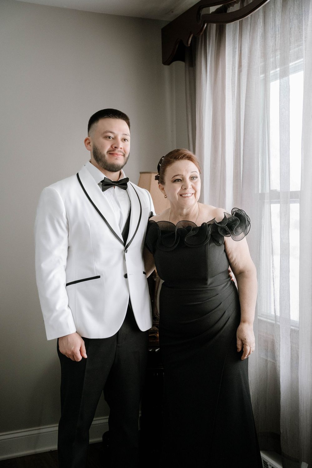 Groom in white tuxedo jacket with bow tie poses with woman in black gown near window.