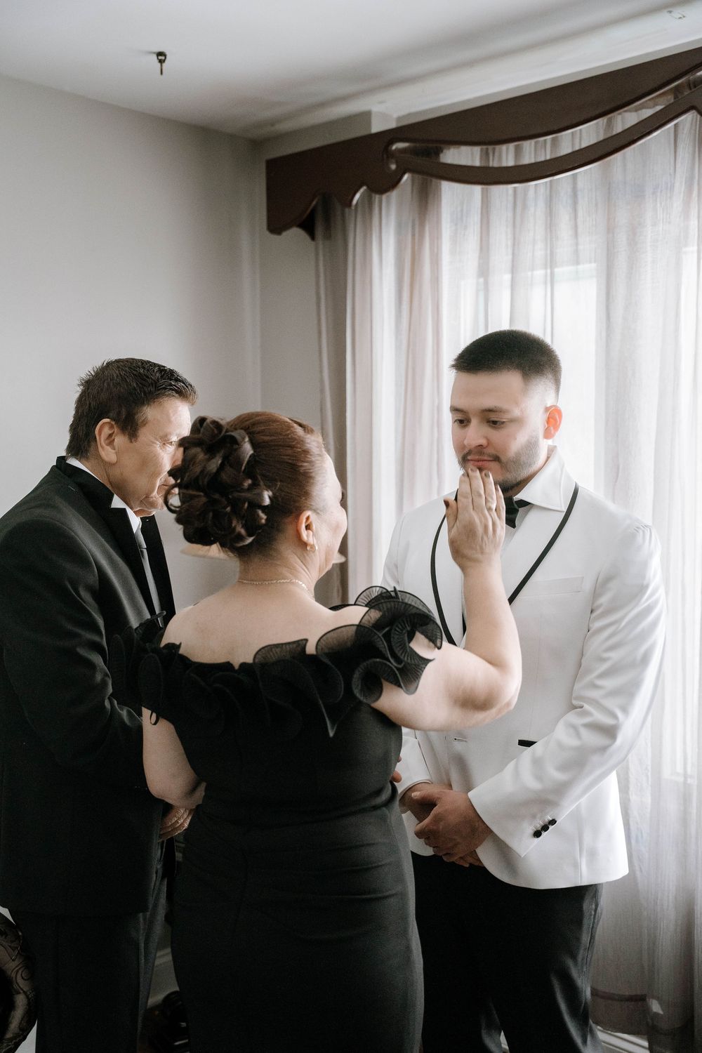 A groom in a white tuxedo receives a tender moment from his parents before his wedding ceremony.